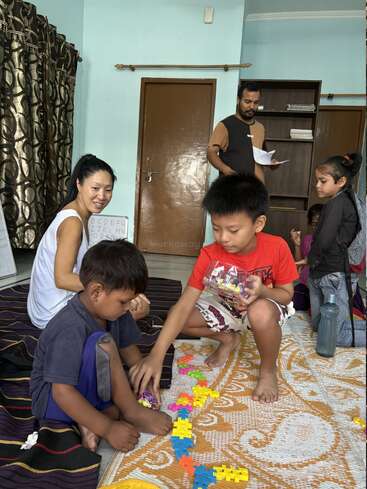 Un grupo de niños y adultos están sentados en el interior, jugando con bloques de construcción de colores en el suelo. Un adulto supervisa mientras los demás participan en el juego creativo.