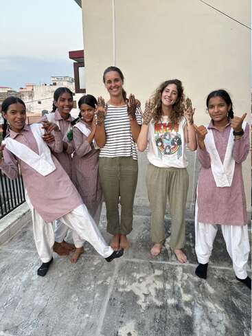 Seis chicas sonrientes están juntas, mostrando sus manos decoradas con henna. Parecen felices y juguetonas, disfrutando de un momento divertido en una azotea a la luz del día.