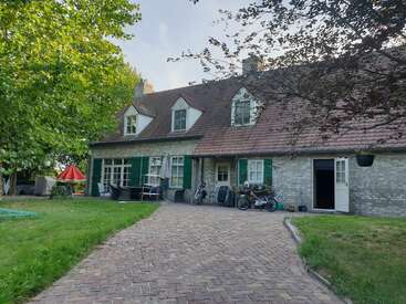 Dieses Bild zeigt ein charmantes Steinhaus mit einem Schrägdach, grünen Fensterläden und einer gemauerten Auffahrt, umgeben von Bäumen, Gras und Gartenmöbeln.