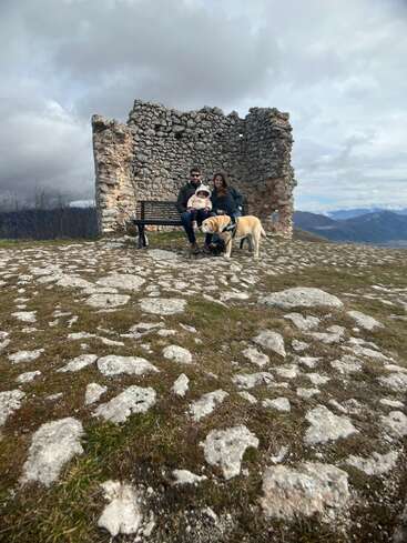 Une famille est assise sur un banc près de ruines de pierre anciennes, accompagnée d'un chien jaune. Le sol rocheux mène à des montagnes lointaines sous un ciel nuageux.