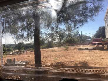 A sunlit outdoor scene viewed through a window, showing a tree, bare dirt ground, scattered pallets, benches, and some building materials against a blue sky.