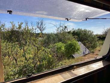 A view through an open window shows green shrubs, sunlight, and a tilted solar panel outside. Blue sky and peaceful, natural surroundings create a serene atmosphere.