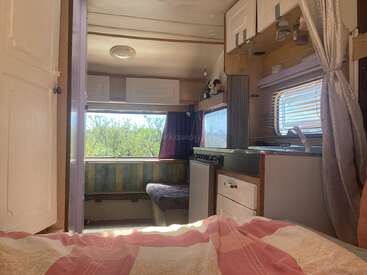 This cozy camper interior features a bed, small kitchen, dining nook with a window, storage cabinets, and natural sunlight streaming in, creating a warm, inviting space.