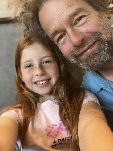 A smiling young girl with long hair takes a close-up selfie with a bearded man. Both appear happy and relaxed, enjoying their time together indoors.