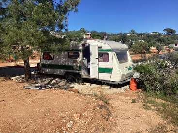 A white caravan with green stripes is parked on rocky ground under a tree. Nearby, an orange gas cylinder and scattered objects are visible. Blue sky.