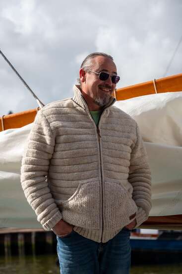 Ein lächelnder Mann mit Sonnenbrille und beiger Strukturjacke steht im Freien, die Hände in den Taschen, neben einem Segelboot mit bedecktem Himmel im Hintergrund.