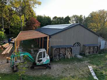 Eine große Holzscheune mit einem Metalldach ist im Bau. Holzstämme sind unter Schuppen gestapelt, ein Minibagger ist geparkt, und Bäume umgeben das Gelände.