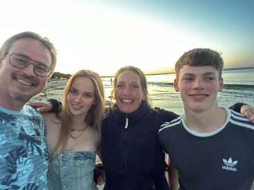 Eine lächelnde Gruppe von vier Personen steht während des Sonnenuntergangs eng zusammen am Strand und genießt die Gesellschaft des anderen mit dem Meer und dem Himmel im Hintergrund.