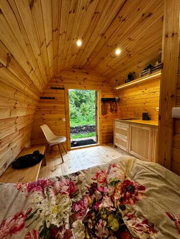 Cette cabane en bois chaleureuse se caractérise par ses murs incurvés, un éclairage tamisé, un lit confortable recouvert d'une parure aux motifs floraux colorés, une chaise simple, des étagères et une vue apaisante sur la verdure à l'extérieur.