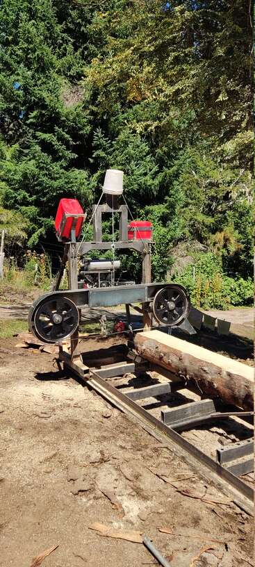 In a forest clearing, a homemade sawmill with large wheels and red containers is cutting a log into lumber, surrounded by trees and sunlight.