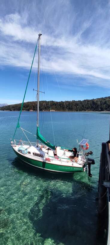 Ein kleines grün-weißes Segelboot liegt auf dem klaren türkisfarbenen Wasser, umgeben von bewaldeten Hügeln, unter einem strahlend blauen Himmel mit Wolkenfetzen.