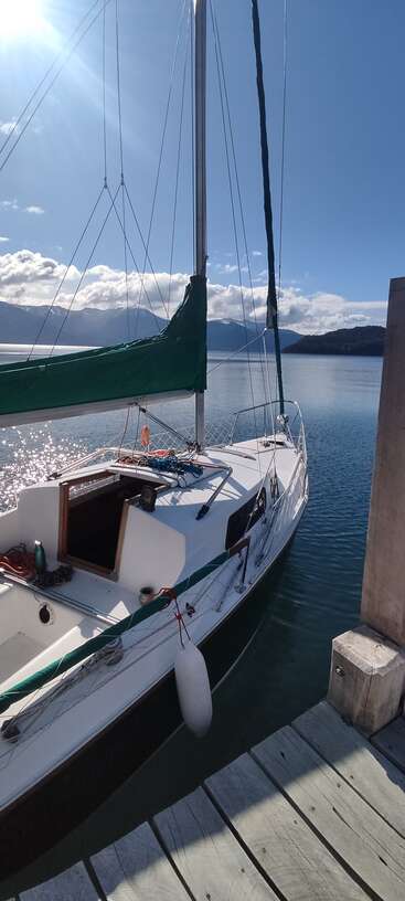 Ein Segelboot mit grüner Plane liegt an einem Holzsteg auf einem friedlichen, sonnigen See, umgeben von schönen Bergen und einem klaren blauen Himmel.