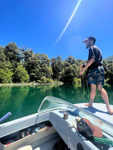 A man stands barefoot on a boat, fishing on a clear lake surrounded by lush trees. Bright sun, blue sky, and relaxed, outdoorsy atmosphere.