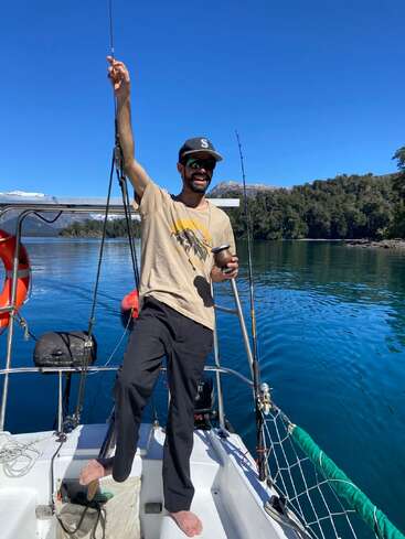 A man stands barefoot on a boat, smiling, holding a drink and fishing rod. Clear blue sky, calm lake, green trees, and distant snowy mountains surround him.