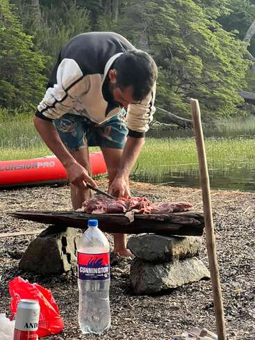 A man prepares raw meat on a rustic wooden slab outdoors, surrounded by forest. Bottled water and a can are placed nearby, creating a camping atmosphere.