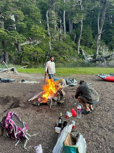 Two people are camping in a forest clearing, tending a blazing campfire surrounded by backpacks, drinks, and supplies, with kayaks and tall trees in the background.