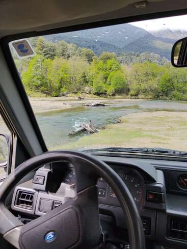 Viewed from inside a vehicle, a scenic landscape reveals a river, driftwood, lush green trees, and distant mountains under a partly cloudy sky, creating a peaceful scene.