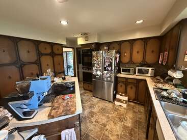 Diese Küche verfügt über dunkle Holzschränke, einen Edelstahlkühlschrank mit Magneten, eine blaue Espressomaschine, Mikrowellen, überfüllte Arbeitsflächen, einen Fliesenboden und Einbauleuchten an der Decke.