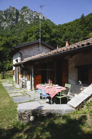 Una acogedora casita de piedra con contraventanas de madera se alza al pie de unas verdes montañas. Fuera, hay una mesa con un mantel a cuadros rojos preparada para desayunar al sol.