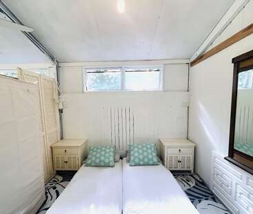 Este quarto aconchegante tem duas camas de solteiro juntas, almofadas com estampas em verde pastel, paredes brancas, mesinhas de cabeceira, um espelho grande, janelas e muita luz natural.