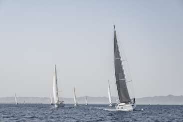 Mehrere Segelboote gleiten unter klarem Himmel über ein ruhiges, blaues Meer. Ferne Berge bilden eine ruhige Kulisse und tragen zur friedlichen und malerischen Atmosphäre bei.