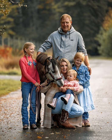 A happy family of five poses outdoors on a path with their small horse. The children smile, one is sitting, surrounded by autumn scenery and trees.
