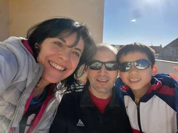 A smiling family of three poses outdoors on a sunny day. The woman, man, and boy wear jackets, and the man and boy wear sunglasses.