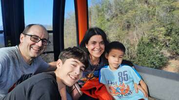 A family of four smiles inside a cable car, enjoying a sunny day. Trees and greenery are visible outside the window, capturing a joyful, memorable moment.