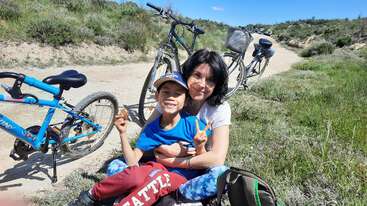 A woman and child sit happily on grass beside two bicycles, both smiling and making peace signs, enjoying a sunny day outdoors on a dirt path.