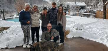 Eine Gruppe von sechs Personen und ein Hund posieren im Winter im Freien. Schnee bedeckt den Boden, ein Pool ist zu sehen und Bäume bilden einen winterlichen, friedlichen Hintergrund.