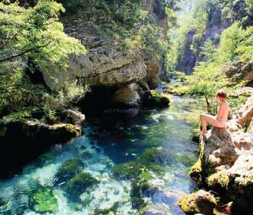 A woman sits on a rock by a river with crystal-clear water, surrounded by lush greenery and large rocks, enjoying the serene natural scenery.