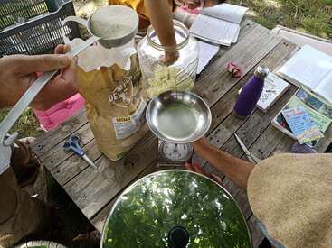 The image depicts a person measuring out flour from a large bag on a wooden picnic table, surrounded by various items and a bowl of green liquid.