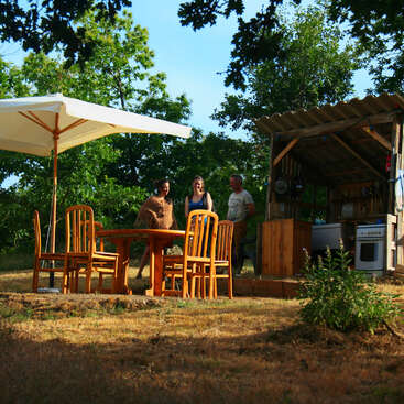 Three people stand near an outdoor wooden kitchen and dining area. Six wooden chairs and a round table sit under a large white umbrella, surrounded by greenery.