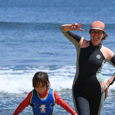 The image depicts a woman and a child on a beach, with the woman wearing a wetsuit and the child wearing a life jacket, both standing near the water\'s edge.