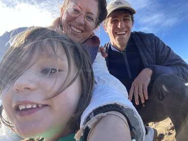Eine fröhliche Familie macht an einem sonnigen Tag am Strand ein Selfie und lächelt, während im Hintergrund der blaue Himmel und der Sand zu sehen sind.