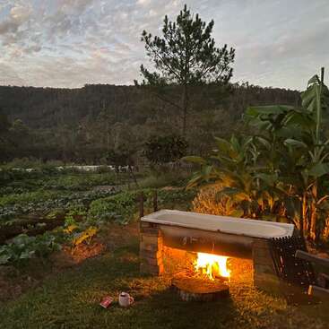 A rustic scene at dusk features a burning fire under a bathtub outdoors, surrounded by lush gardens, trees, mountains, and a cup placed on the grass.