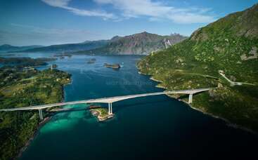 An elegant bridge stretches across deep blue waters, connecting lush green hills and islands, surrounded by majestic mountains under a vibrant blue sky with scattered clouds.