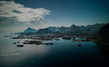The image shows a coastal landscape with striking mountains, small islands, and calm blue water. A town nestles at the foot of the dramatic, rugged peaks.