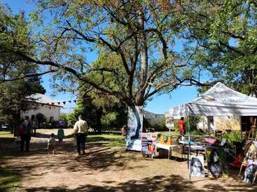 Uma cena ensolarada de um mercado ao ar livre mostra pessoas caminhando sob uma grande árvore. Uma barraca de arte com retratos é montada sob um dossel branco ao lado de um caminho.