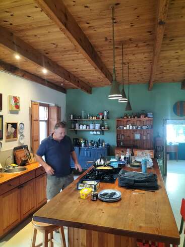 Um homem está cozinhando em uma cozinha rústica com armários de madeira, vigas expostas e luzes pendentes. A cozinha é aconchegante, colorida e repleta de vários utensílios.