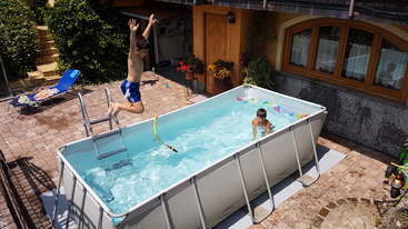 The image shows a backyard with a pool and a fence, featuring a large tree in the background, with a house visible in the distance. The scene is set against a natural, outdoor backdrop.
