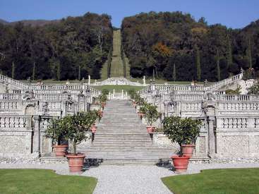 The image depicts a grand stone staircase with ornate railings, flanked by potted trees and lush greenery, set against a serene blue sky backdrop.