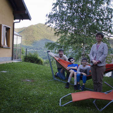 The image depicts a family of four posing for a photo in a backyard, with a hammock and lounge chair on the grass, set against a mountainous backdrop.