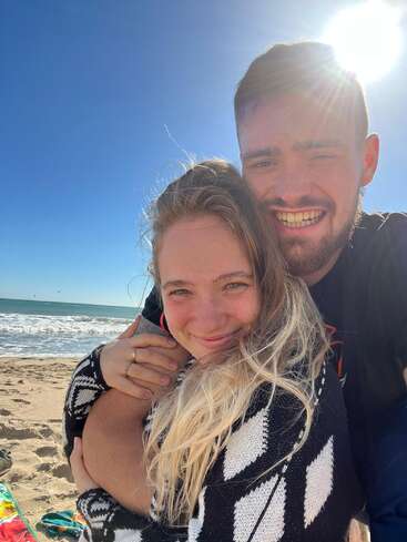 Das Bild zeigt ein Paar, das sich an einem sonnenverwöhnten Strand umarmt, mit dem Arm des Mannes um die Schultern der Frau, vor einer ruhigen Meereskulisse.