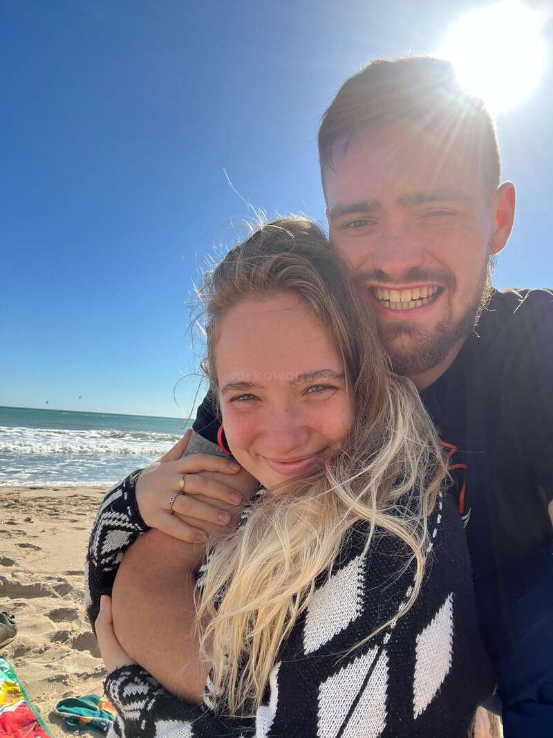 Das Bild zeigt ein Paar, das sich an einem sonnenverwöhnten Strand umarmt, mit dem Arm des Mannes um die Schultern der Frau, vor einer ruhigen Meereskulisse.