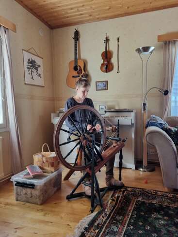 Uma pessoa está sentada dentro de casa, usando uma roda de fiar. Instrumentos musicais estão pendurados na parede. Um ambiente aconchegante com teto de madeira, tapetes e luz natural entrando pela janela.