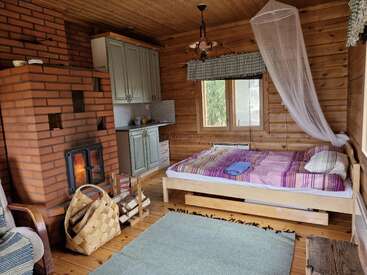 Cette chambre confortable, aménagée dans une cabane en bois, dispose d'un lit, d'une cheminée, d'une kitchenette, d'une fenêtre et d'une décoration rustique. Du bois de chauffage et un panier se trouvent près du poêle. L'ambiance y est chaleureuse et conviviale.