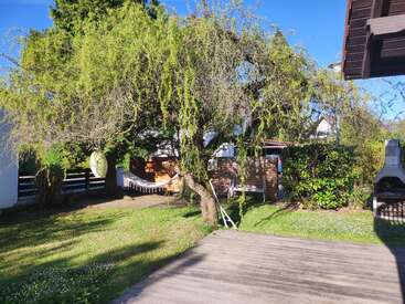Ein sonniger Garten verfügt über einen großen Baum mit einer hängenden Laterne, eine Hängematte im Schatten, grünen Rasen, eine Holzterrasse, üppige Büsche und einen Außengrill.
