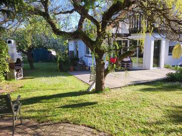 Ein sonniger Garten mit einem großen, grünen Baum, einer Holzterrasse, einer Rasenfläche, Gartenmöbeln, einem Grill und einem gemütlichen Haus mit Glastüren.