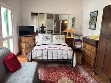 A cozy bedroom features a wrought iron bed, floral pillows, antique furniture, a red patterned rug, a TV, mirrors, wooden drawers, and warm ambient lighting.